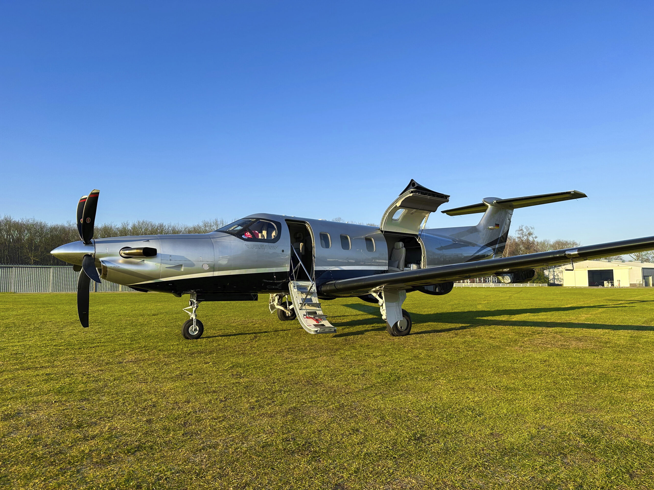 Pilatus PC-12 NGX – Access to Special Destinations A Lohn Aviation Group Pilatus PC-12 NGX on a grass runway with its cabin door and cargo door open.